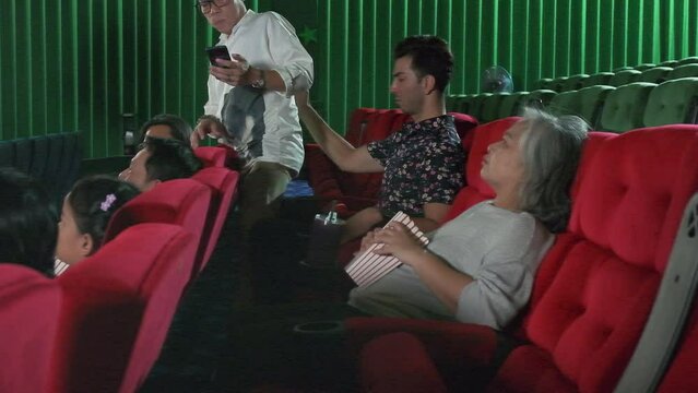 Late man walks to movie theater seat, uses mobile phone, interrupts Latin man, senior woman, and another audience, has bad manners behavior when watching cinema films, and is annoyed with discipline.