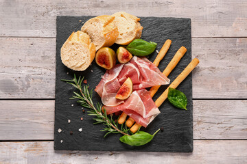 Board with slices of tasty prosciutto, basil, bread, Italian Grissini and figs on grey wooden...