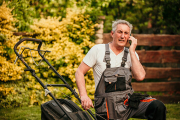 Elderly gentleman in a suit standing beside a malfunctioning lawnmower, seeking help via his smartphone