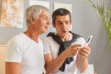 Shocked adult man sitting with his senior father in living room at home using smartphone see shock...