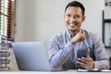 Fototapeta na wymiar Portrait of a cheerful man using smart phone at home office. freelance asian man entrepreneur and working from home research, planning and info 