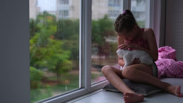 A little girl sits by the window and pets a kitten. The child sadly looks out the window at the rain.
