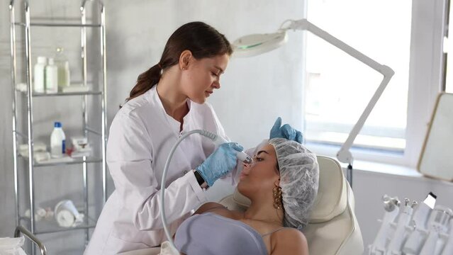 Positive cosmetologist performing machine-based facial procedure on young female client employing ion bubble pen attachment in cosmetic clinic, aimed at gentle cleanse and hydration of skin