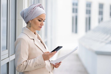 Businesswoman in formal jacket looks at phone screen holding papers. Concentrated Muslim woman messages colleague about important work project