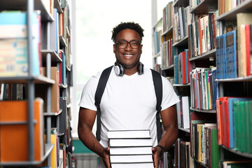 Cheerful african male student in fashionable clothes with stack of books in university library....