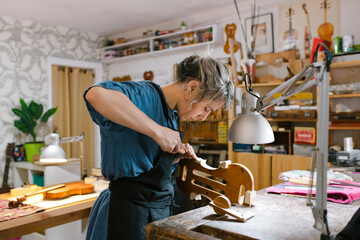 Luthier with hand tool carving on violin at desk in workshop