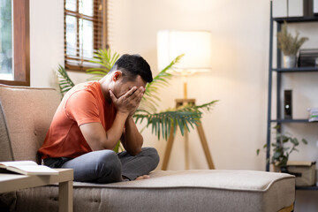 Sad bearded middle-aged man in casual sitting on couch at home, leaning on hands and looking down,...