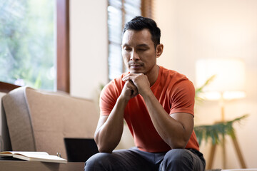 Sad bearded middle-aged man in casual sitting on couch at home, leaning on hands and looking down,...
