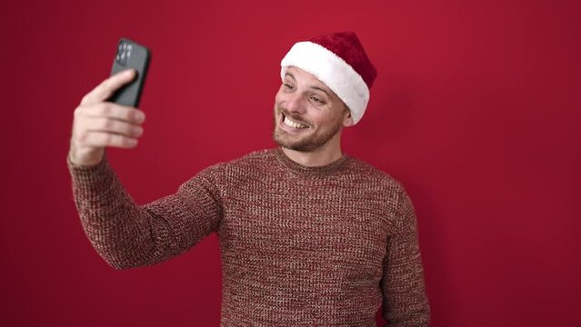 Young caucasian man smiling taking selfie picture wearing christmas hat over isolated red background