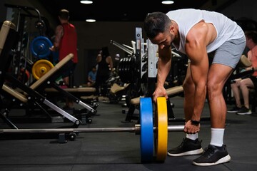 Fototapeta na wymiar Strong Hispanic man putting weight plate on barbell before deadlift at a gym.