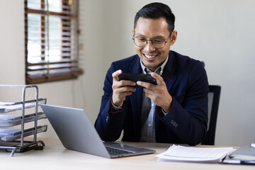 Fototapeta na wymiar Texting to colleague or play games. Confident young asian man in smart casual wear holding smart phone and looking at it while sitting at his working place in office