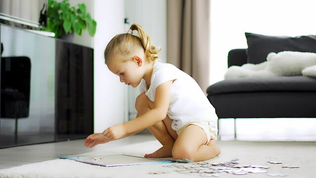 Little blonde girl sits at home on the carpet and collects puzzles