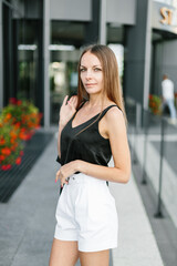 A girl with long blond hair, wearing a black top, black jacket and white shorts, poses against the backdrop of modern architecture. Young woman walking in a park with landscape design