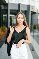 A girl with long blond hair, wearing a black top, black jacket and white shorts, poses against the backdrop of modern architecture. Young woman walking in a park with landscape design