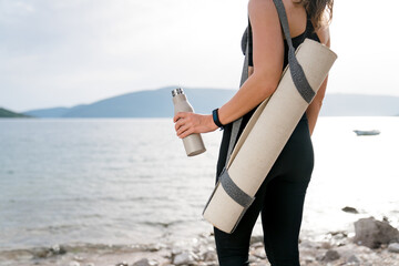 Yoga mat, reusable water bottle in woman hand. Sport, wellness on sea beach. Girl resting after workout outdoor, fitness in nature. Concept of healthy, sustainable lifestyle, ecological responsibility