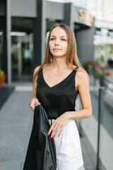 A girl with long blond hair, wearing a black top, black jacket and white shorts, poses against the backdrop of modern architecture. Young woman walking in a park with landscape design