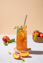 Refreshing peach ice and mint tea. Vegan homemade cold summer drink on tall glass on a light background with fresh fruits and shadows.