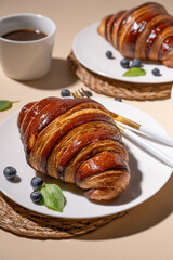 Freshly baked croissants with chocolate on a white plate and blueberries on a yellow background with cup of black coffee close up.