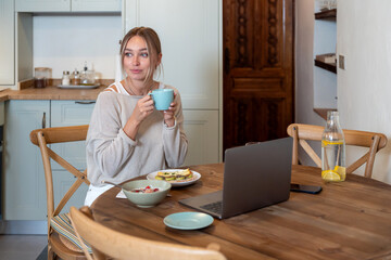 Caucasian female eating delicious breakfast with fresh strawberries while sitting on kitchen and working on her laptop