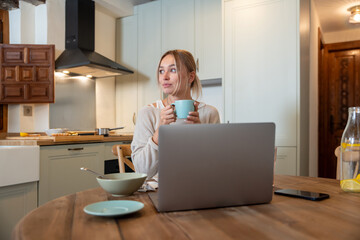 Caucasian female eating delicious breakfast while sitting on kitchen and working on her laptop