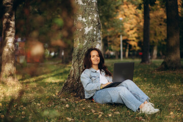 Woman sitting under the tree in park working on laptop and drinking coffee