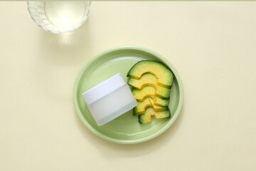 Minimalist scene for advertising cosmetic, cream or mask of natural extract. A cosmetic jar with slices of avocado displayed on round ceramic dish on beige background with glass cup