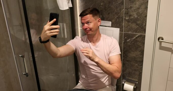 Man sitting on toilet and taking selfie on his mobile phone camera 4k movie. When you are bored at home concept 