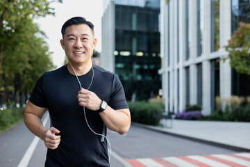 Portrait of an Asian young man running in headphones on a city street in the morning, smiling at...