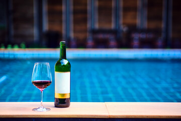 A bottle of red wine and a glass of wine are set on the edge of a clear blue pool, surrounded by a lush green lawn. The afternoon sun shines down, creating a relaxing and inviting atmosphere. - Powered by Adobe