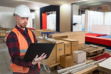 Worker with laptop. Man in furniture workshop. Manufacturing specialist. Wooden boxes and pallets are behind man. Worker in orange vest and helmet. Furniture workshop contractor. Engineer, foreman
