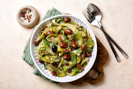 Italian salad made with sauteed broad-leaved endive or escarole with taggiasca olives, pine nuts, raisins. Vegan dish. Top view.