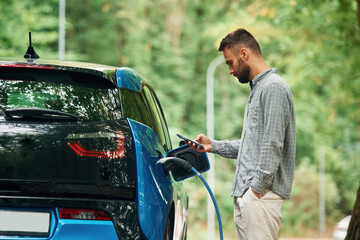 Handsome man is standing outdoors near electric automobile and waiting for charging