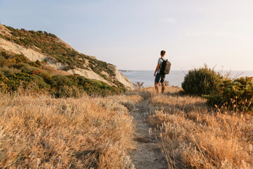 Hiker man in a T-shirt and shorts with backpack on the coast with warm sunlight.