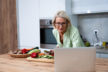 Senior adult woman cooking healthy food for dinner in her domestic kitchen using laptop computer to read online recipe. Grandmother preparing vegetables for guests to eat. - obrazy, fototapety, plakaty