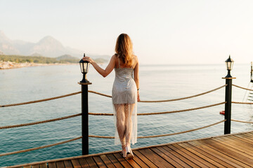 A slender girl with long hair in a gray shiny dress walks along the pier by the sea at sunset. A...