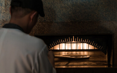 Chef in uniform holding an floured metal paddle for cooking pizza into a fired oven. The process of making pizza. Selective focus to pizza in oven.
