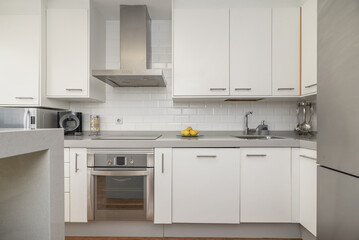 Front image with a kitchen equipped with white furniture, integrated stainless steel appliances, a small island on one side, small appliances on a light gray countertop and wooden floor