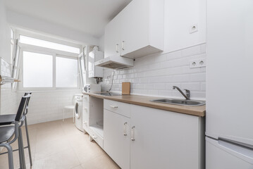 Furnished kitchen on one of the walls with white furniture, integrated white appliances, high stools on one side and a light wood-colored countertop and a white aluminum window