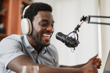 Young african man host recording podcast inside studio at home using headphones, microphone and...