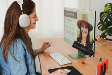Mature female influencer recording a podcast inside modern office using microphone, headphone and  computer while streaming live with mobile phone