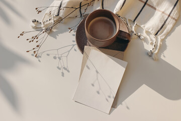 Christmas still life. Blank greeting card, invitation mockup with dry flower in sunlight. Soft shadows. Cup of coffee and white checkered plaid on table. Winter flatlay, top view. Copy space.