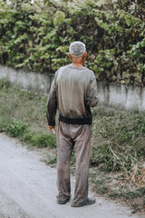An old adult gray-haired thin homeless hungry man walks along the street road in search of food....