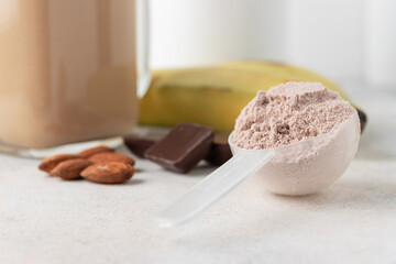 Chocolate whey protein powder in measuring spoon, glass jar of protein milkshake drink or smoothie, bananas, chocolate cubes and almonds on white background. sport nutrition, food supplements