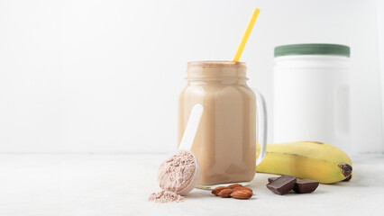 Chocolate whey protein powder in measuring spoon, glass jar of protein milkshake drink or smoothie, bananas, chocolate cubes and almonds on white background. sport nutrition, food supplements