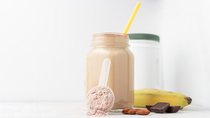 Chocolate whey protein powder in measuring spoon, glass jar of protein milkshake drink or smoothie, bananas, chocolate cubes and almonds on white background. sport nutrition, food supplements