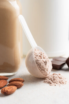 Chocolate Whey Protein Powder In Measuring Spoon, Glass Jar Of Protein Milkshake Drink Or Smoothie, Chocolate Cubes And Almonds On White Background. Sport Nutrition, Food Supplements