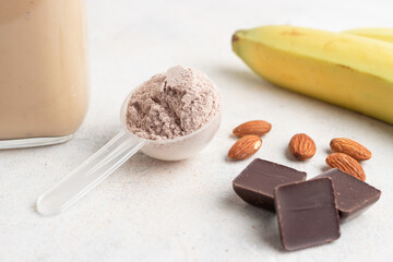 Chocolate whey protein powder in measuring spoon, glass jar of protein milkshake drink or smoothie, bananas, chocolate cubes and almonds on white background. sport nutrition, food supplements