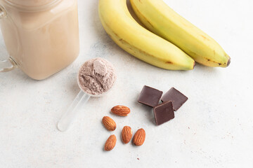 Chocolate whey protein powder in measuring spoon, glass jar of protein milkshake drink or smoothie, bananas, chocolate cubes and almonds on white background. sport nutrition, food supplements