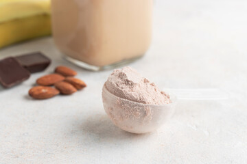 Chocolate whey protein powder in measuring spoon, glass jar of protein milkshake drink or smoothie, bananas, chocolate cubes and almonds on white background. sport nutrition, food supplements