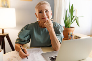 Pretty elderly bossy stylish female HR manager working at her office in front of laptop, signing papers or reading CV of new candidate on vacant position in their company, looking at camera smiling - 659511638